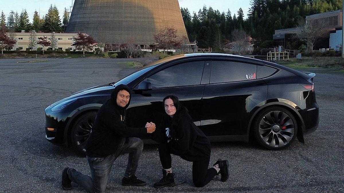 Eddi and Suzie posing beside their black Model Y in a parking lot near an industrial cooling tower.