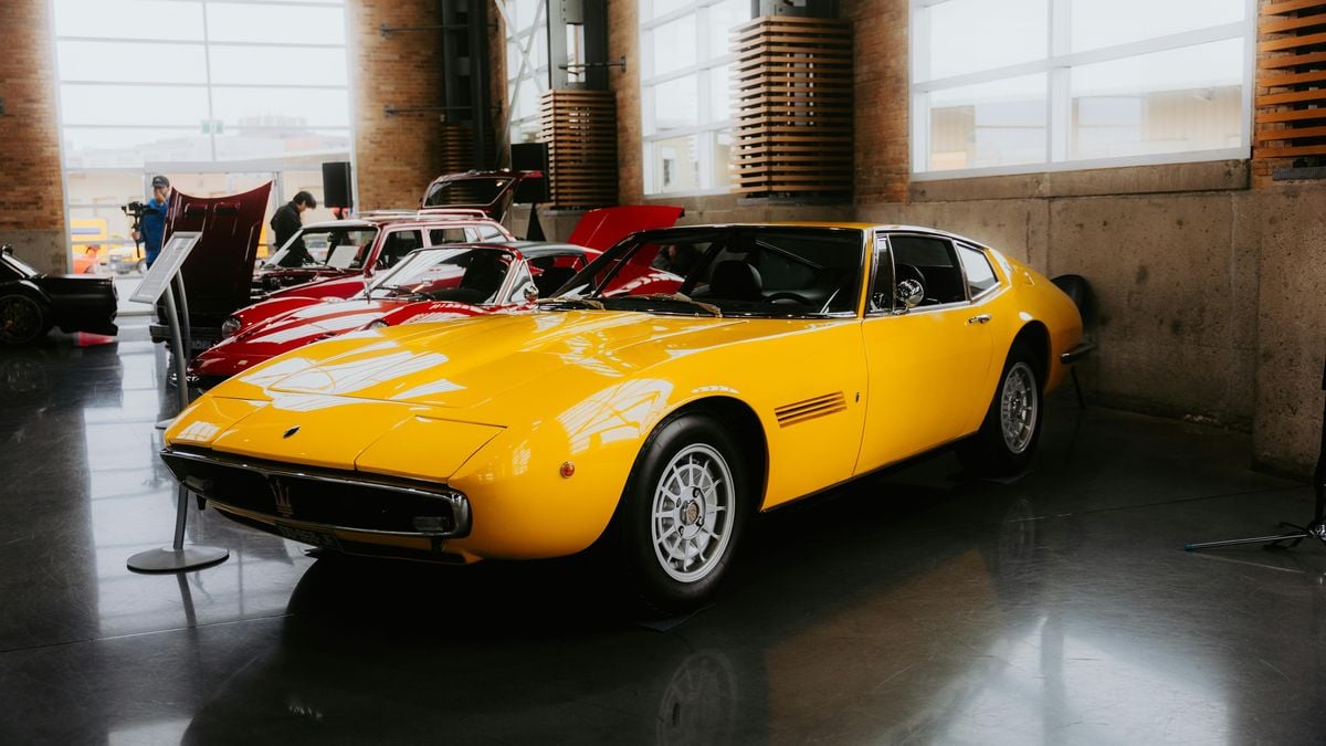 A vintage yellow Maserati sports car on display in a well-lit exhibition hall, surrounded by other classic cars.