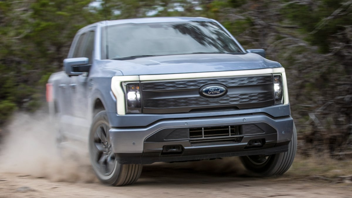 Ford F-150 Lightning electric pickup truck driving fast on dirt road with dust trail in forest setting