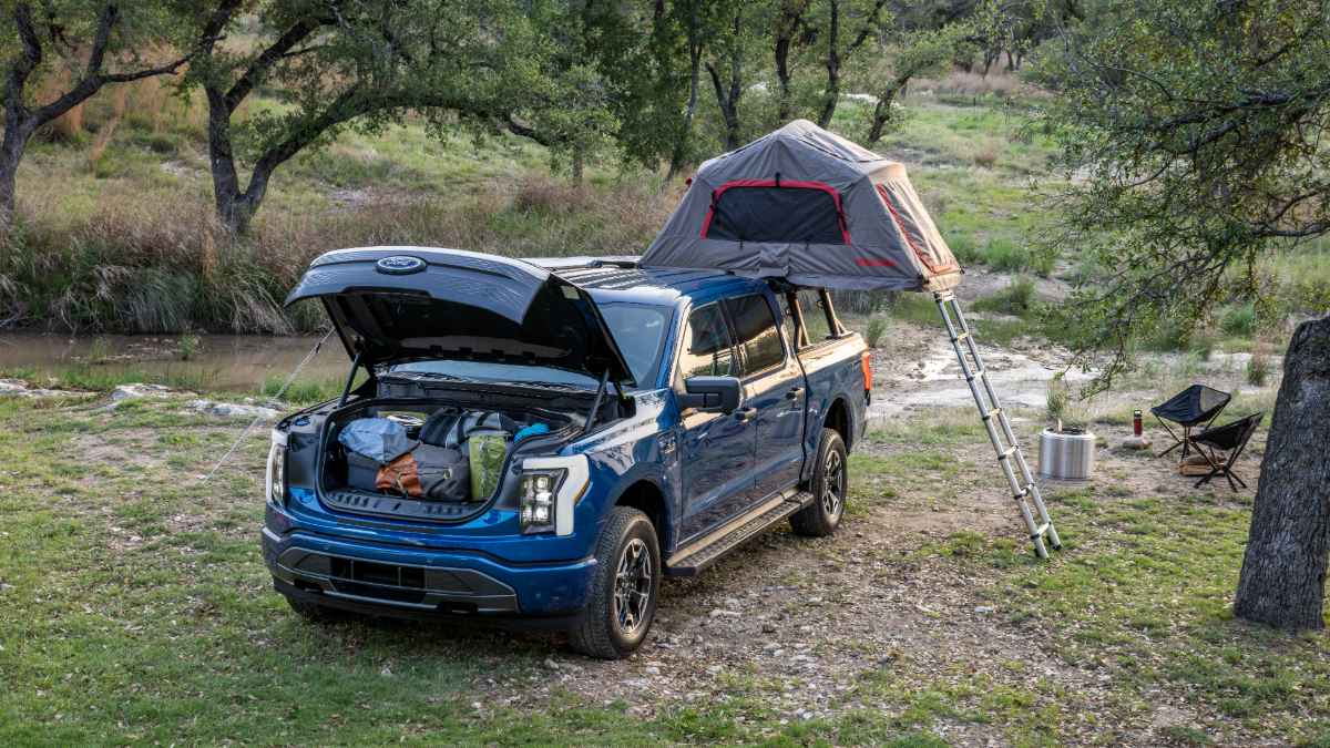 Blue Ford F-150 Lightning electric truck front three-quarter view parked outdoors with rooftop tent and camping gear