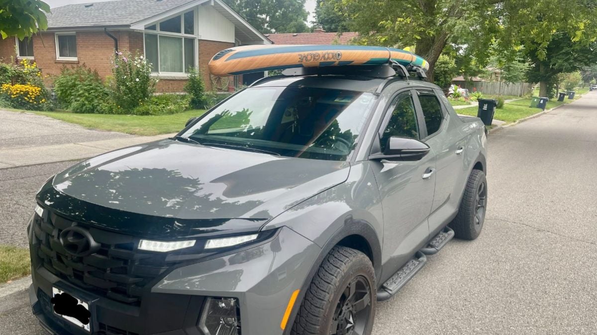 Gray Hyundai Santa Cruz with a paddleboard secured on the roof, parked on a residential street. Green trees and a brick house with a garden are in the background.