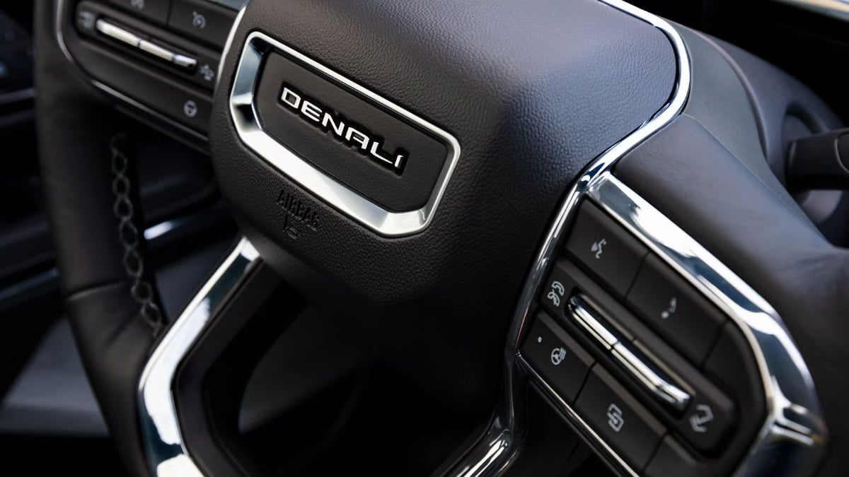 Close-up interior detail of a GMC Sierra EV Denali dashboard controls, showing black textured surface with chrome-trimmed buttons and distinctive Denali branding.