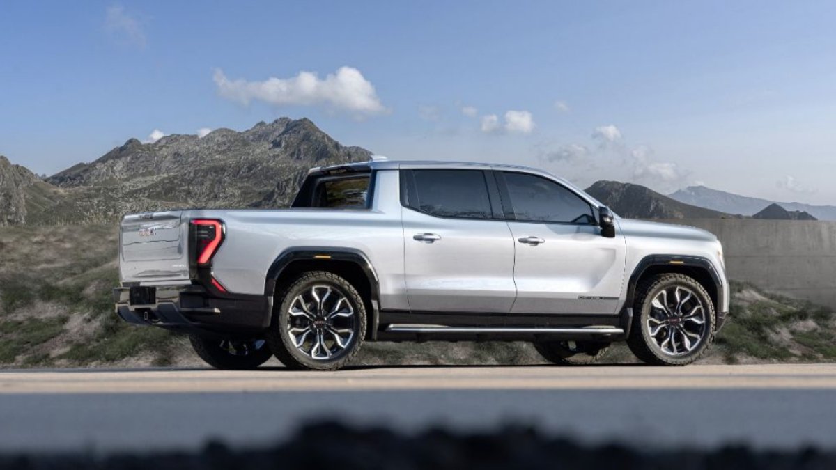 White GMC Sierra pickup truck parked on mountain road with rugged peaks and blue sky backdrop, side profile view with stylish alloy wheels