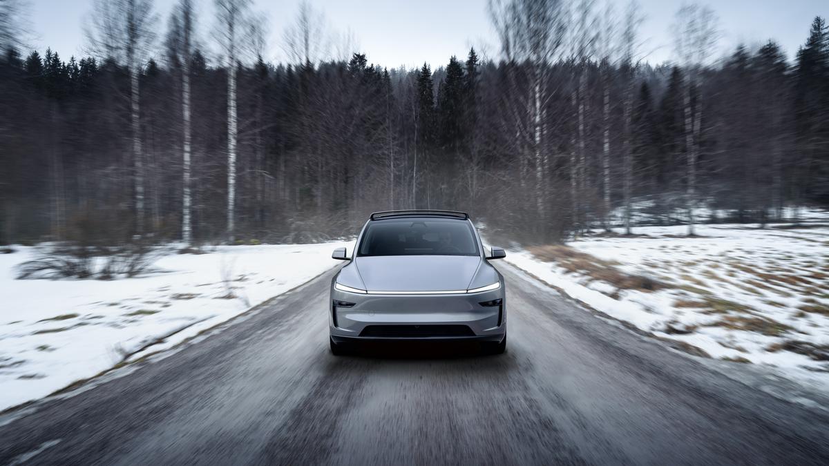 A sleek silver electric car drives down a snowy road, flanked by bare trees under a gray sky.
