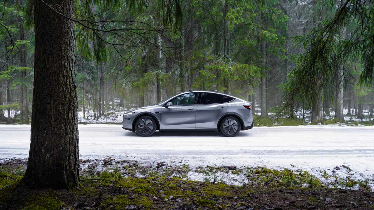 A sleek silver electric car drives on a snowy road, surrounded by tall green trees in a serene forest setting.