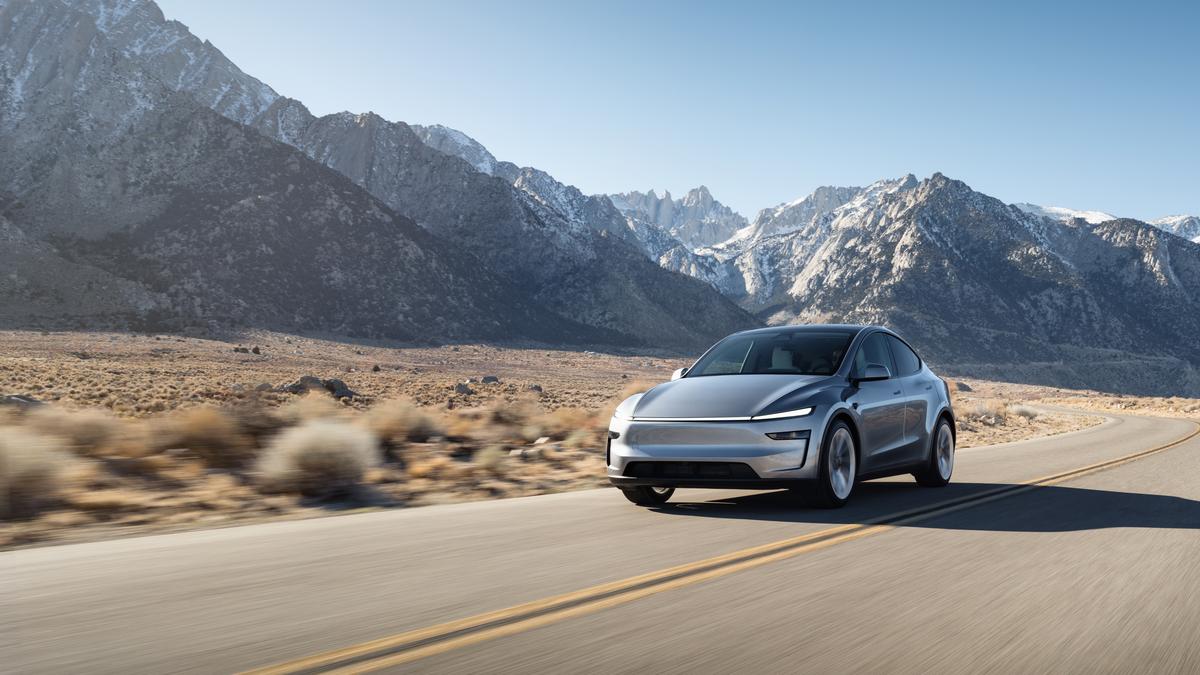 A 2025 Gray Tesla Model Y driving along a winding road with majestic snow-capped mountains in the background. Clear blue sky above.