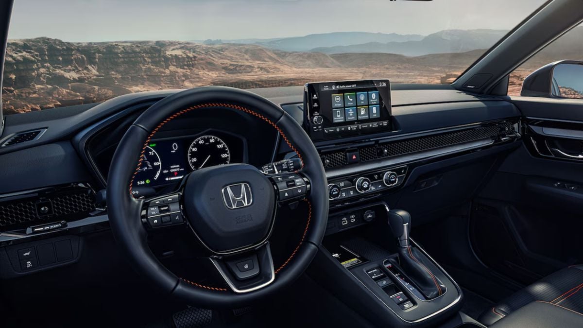Honda CR-V interior featuring a black steering wheel with orange stitching, a digital dashboard, and a central touchscreen. A scenic desert landscape is visible through the windshield.