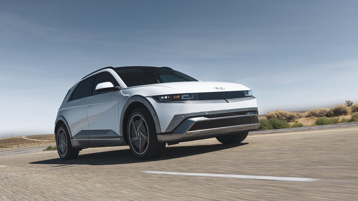 A sleek white Hyundai Ioniq 5 electric vehicle driving on a sunny, open road with a clear blue sky in the background.