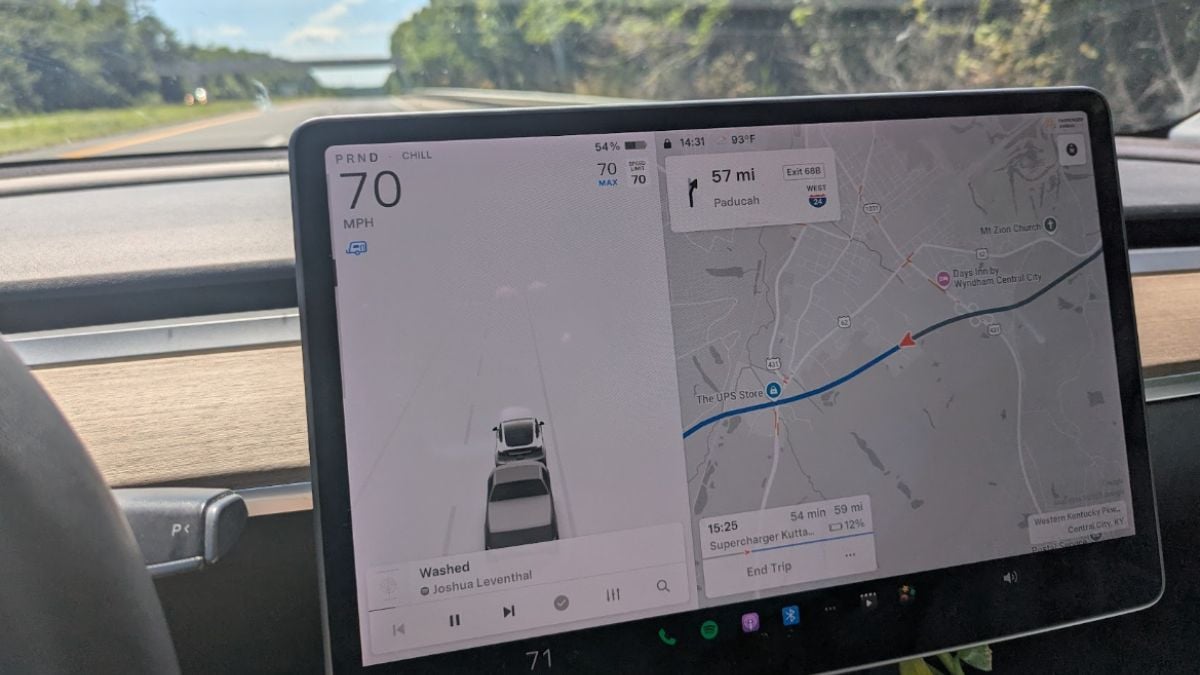 Close-up of a Model Y's dashboard screen showing navigation and driving data. The car is traveling at 70 mph with a map indicating 57 miles to the destination. Sunlight illuminates the road ahead, lined with greenery, suggesting a calm drive.