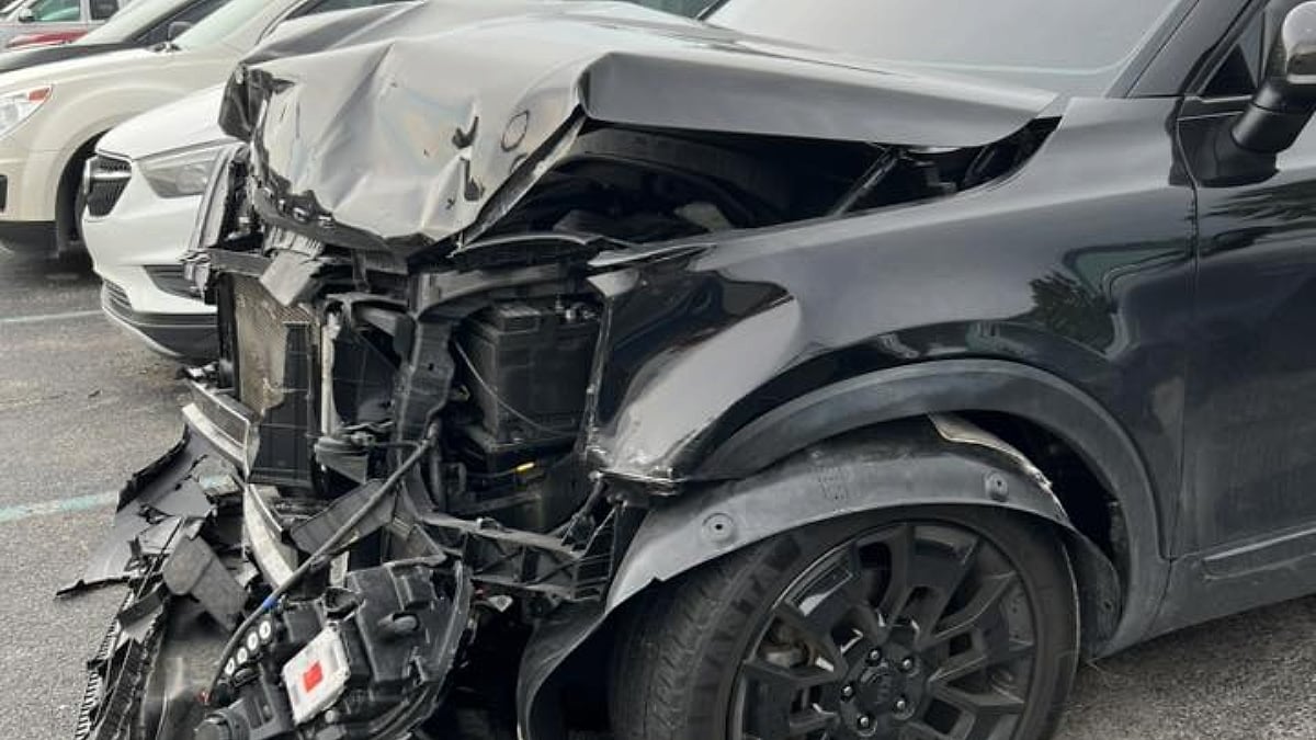 Lacie's black Kia Telluride with severe front-end damage parked in a lot. The hood is crumpled, exposing internal components. Nearby are other undamaged cars.