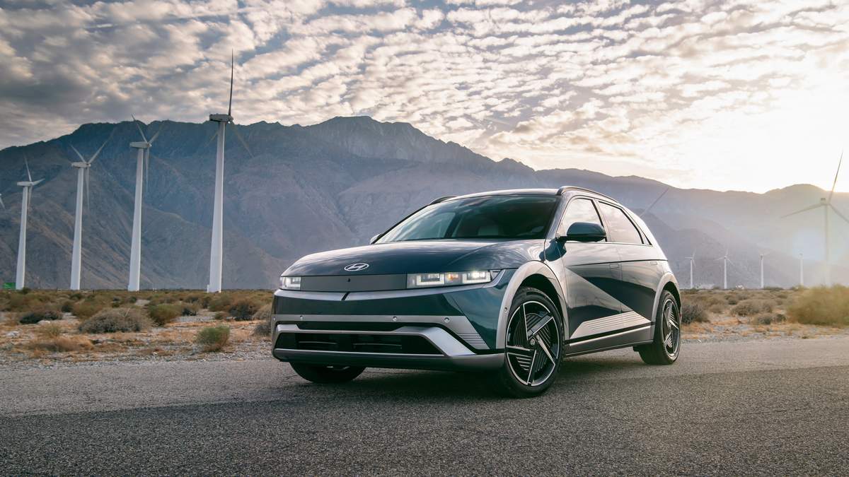 A sleek Hyundai Ioniq 5 parked on a desert road, with wind turbines and mountains in the background under a cloudy sky.