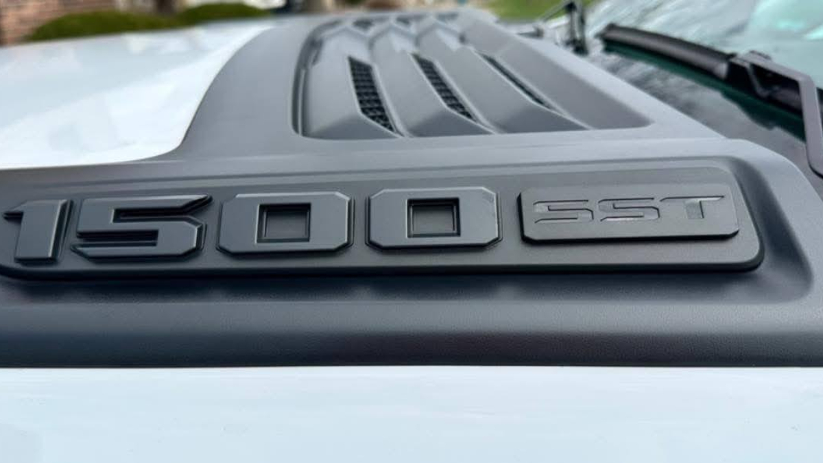 Close-up of a new Ram 1500's hood featuring bold black text "1500 SST" on a sleek, raised, and vented design. The setting conveys a sense of strength and modern style.