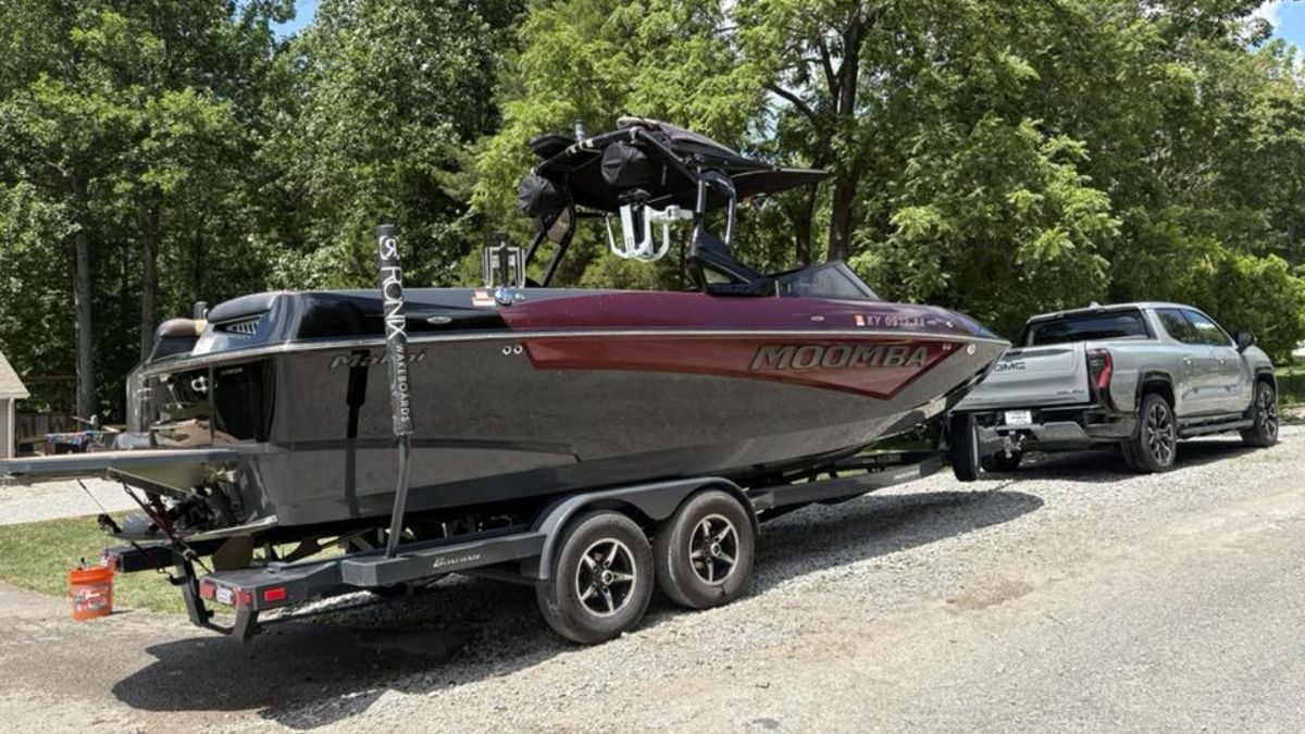 A sleek black and red Moomba boat on a trailer is hitched to a silver GMC Sierra EV, parked on a gravel path amidst lush green trees under a clear sky.
