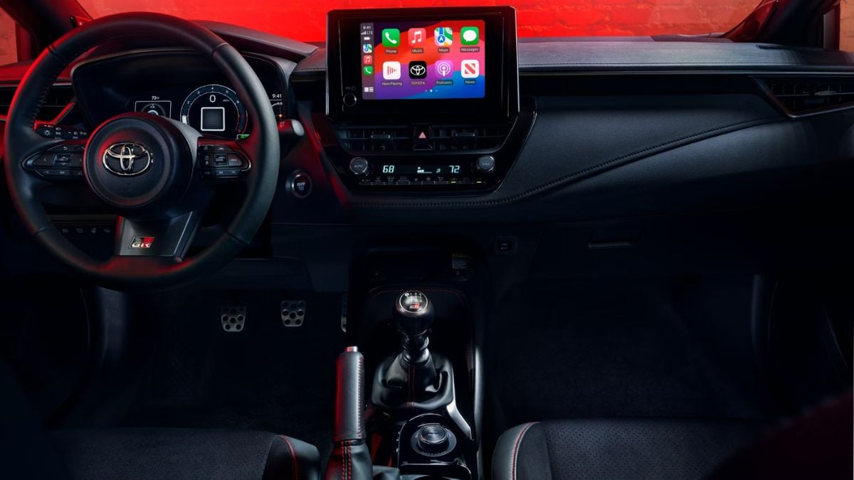 Toyota GR Corolla interior with a sporty aesthetic, showcasing a steering wheel with an emblem, manual gear stick, and a central touchscreen display amid soft red lighting.