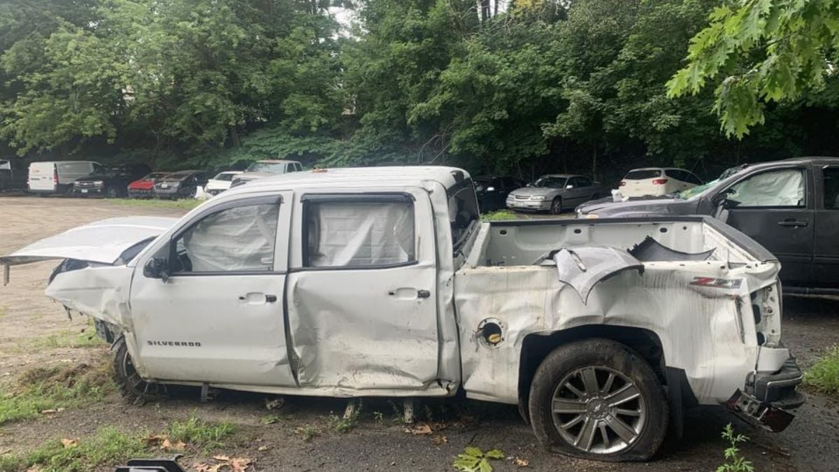 A severely damaged white Chevrolet Silverado with missing rear wheel and crushed bed. The vehicle is parked on dirt, surrounded by trees and other parked cars.