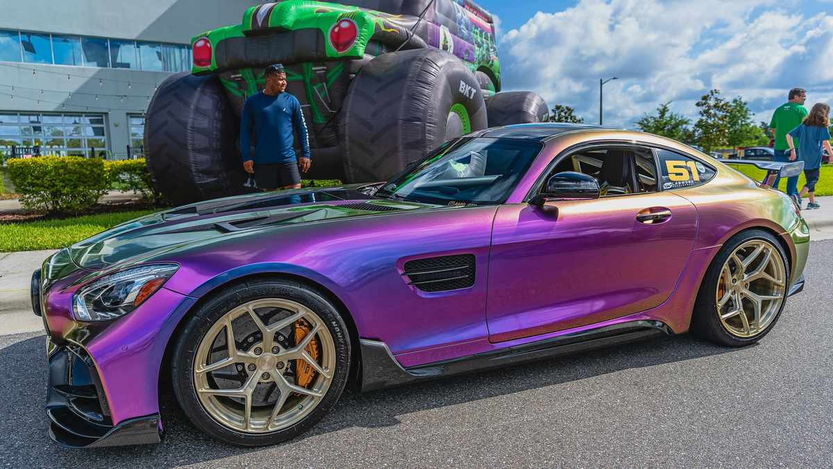 A vibrant, iridescent purple and green sports car showcases its sleek design and gold rims, parked near a large monster truck inflatable.