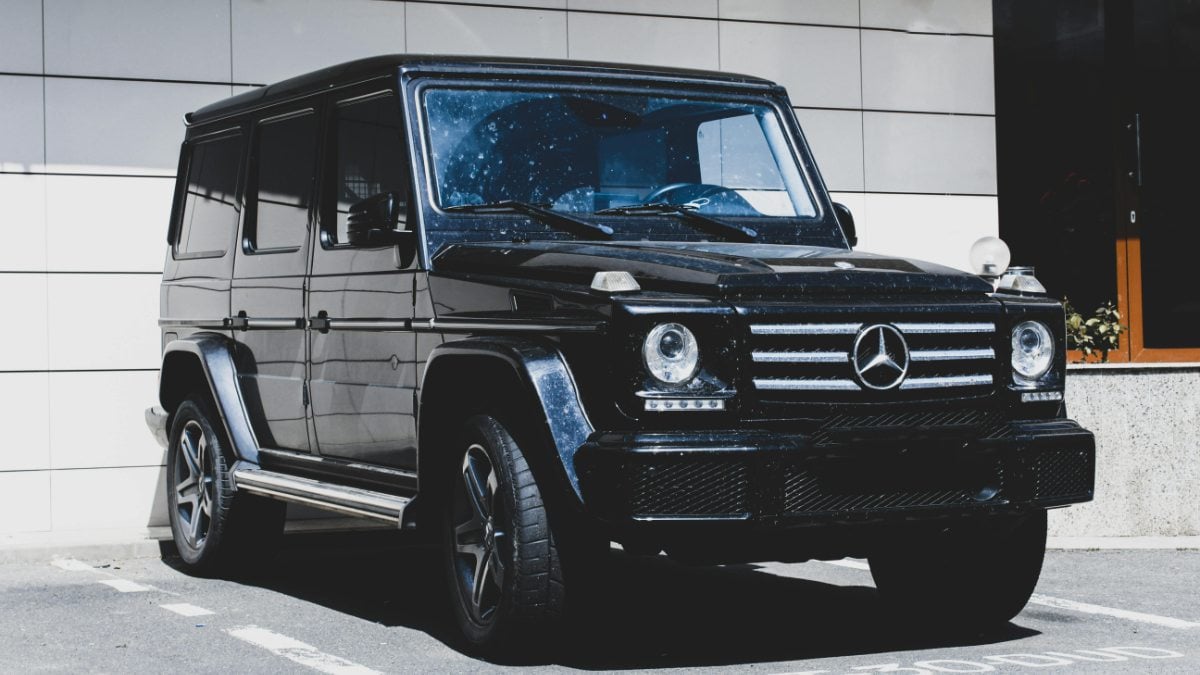 A black Mercedes-Benz G-Class SUV shown from the front three-quarter angle, featuring LED daytime running lights and the distinctive boxy styling of the G-Wagon, parked against a white building exterior.