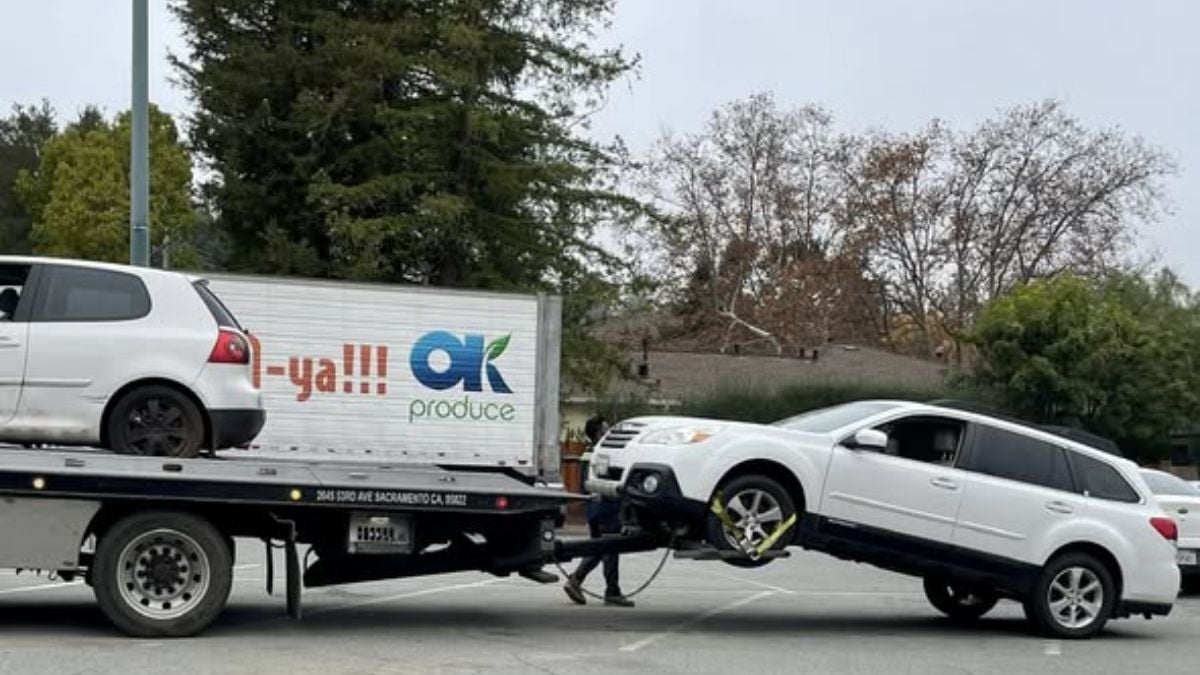 A white Subaru being loaded onto a flatbed tow truck alongside another white vehicle, with an "OK Produce" delivery truck visible in the background.