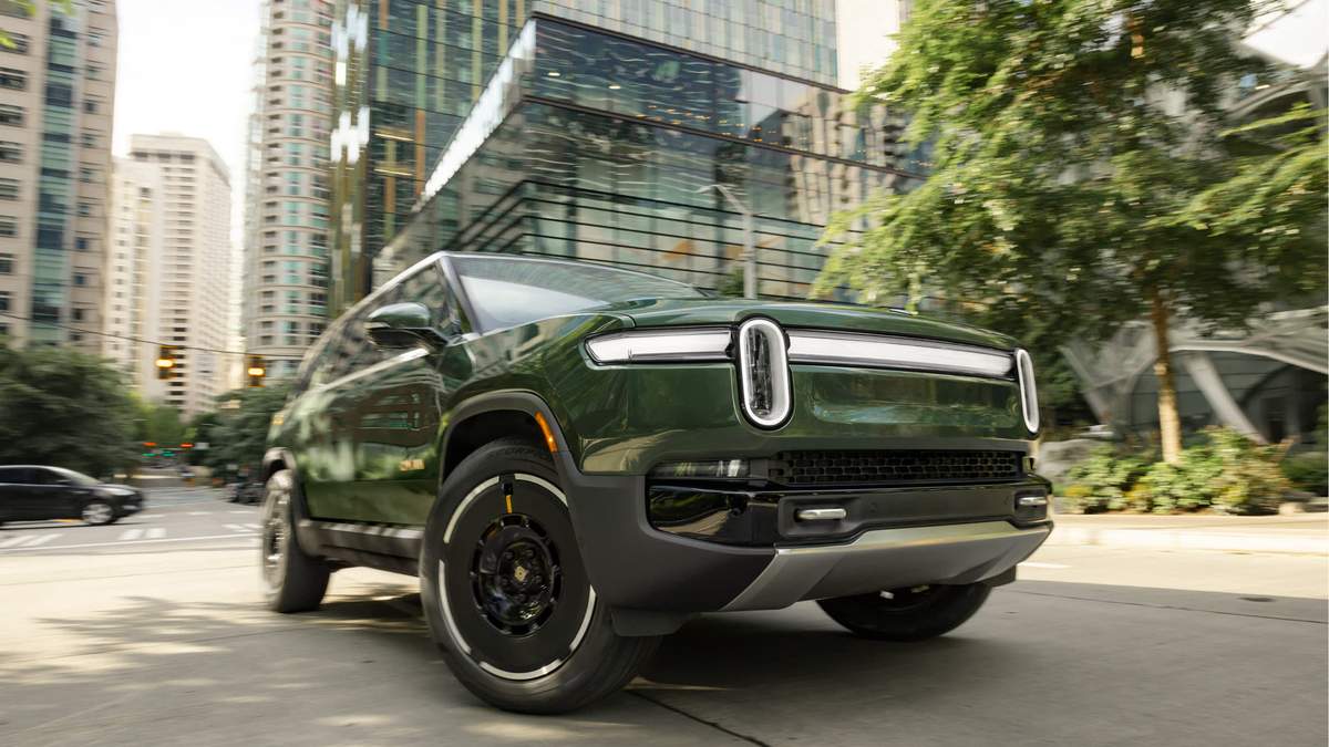Dark green Rivian R1S electric SUV parked on urban street with modern glass buildings and trees in background