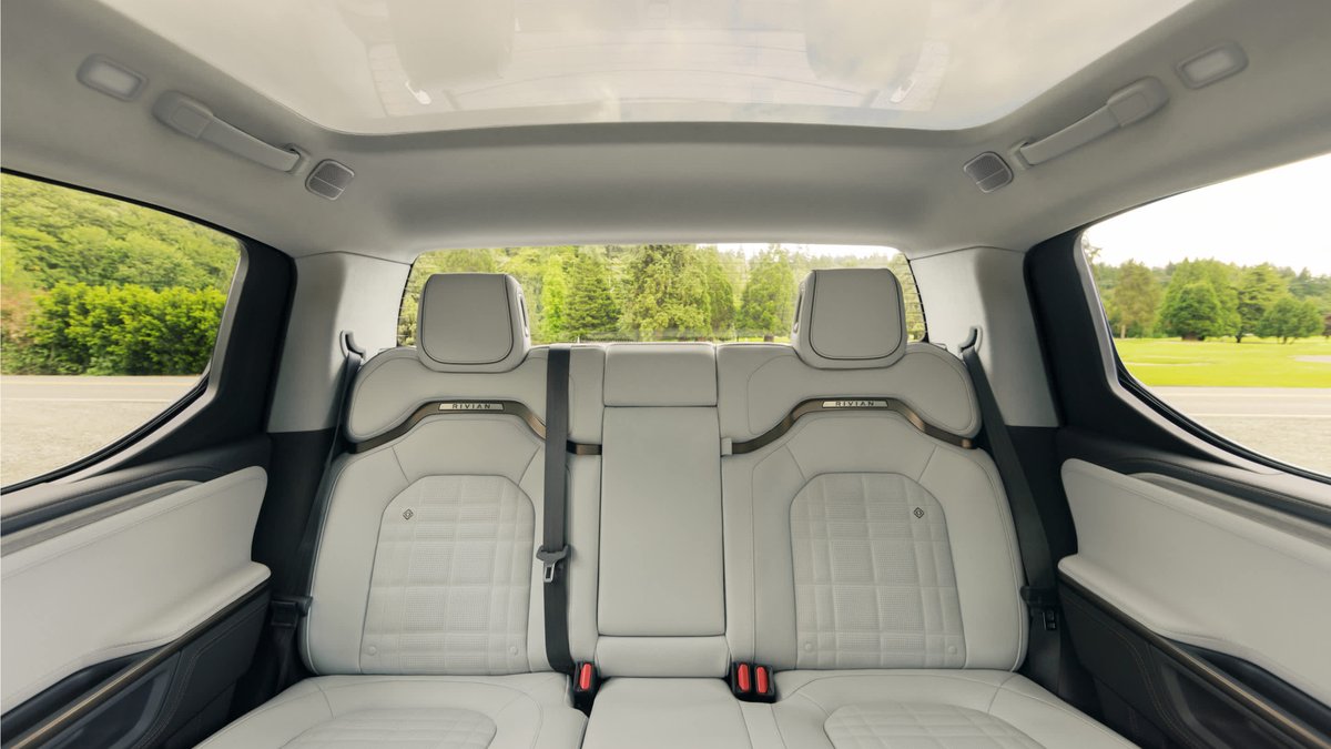 Interior view of a Rivian R1T with leather seats, panoramic sunroof, and spacious rear seating, surrounded by lush greenery outside.