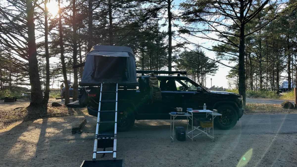 Black 2023 Rivian R1T side view with a tent on top, parked at a campsite with a ladder and camping setup nearby.