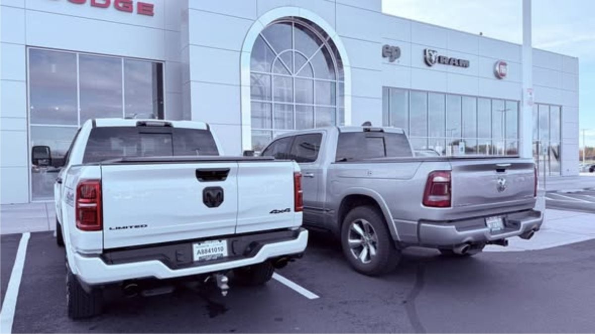 Two Ram 1500's, one white and one gray, are parked outside a car dealership with large windows and signs. The mood is calm and commercial.