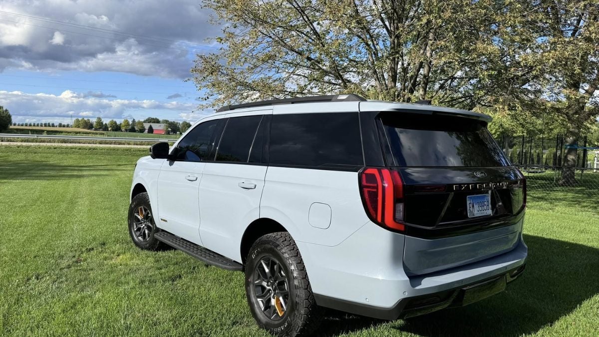 Grey SUV parked on lush green grass under a clear sky with scattered clouds. Tall trees in the background convey a serene, peaceful atmosphere.
