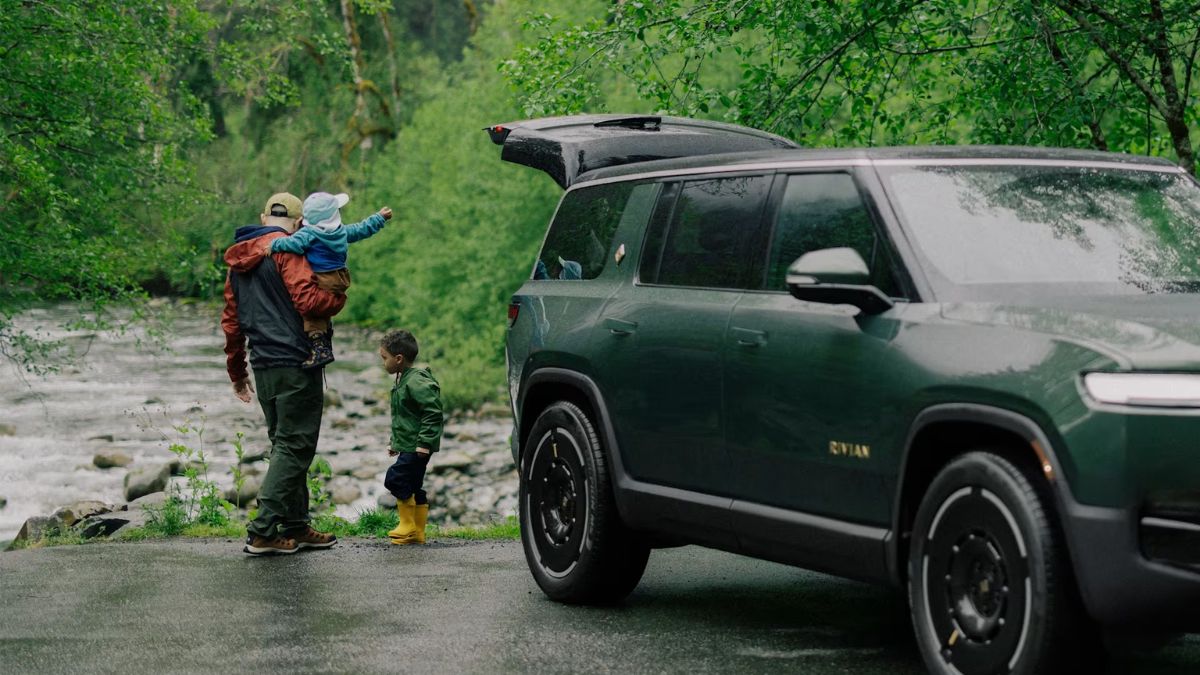 A parent holds a child by a riverside, near a green Rivian R1T with an open trunk. A toddler in yellow boots stands nearby. The scene is lush and serene.
