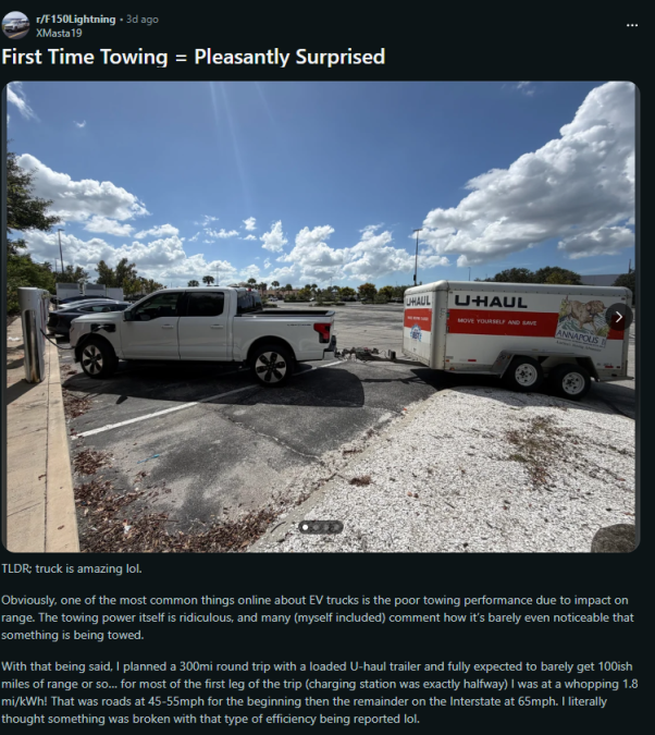A white electric truck is towing a U-Haul trailer in a parking lot under a blue sky with scattered clouds.