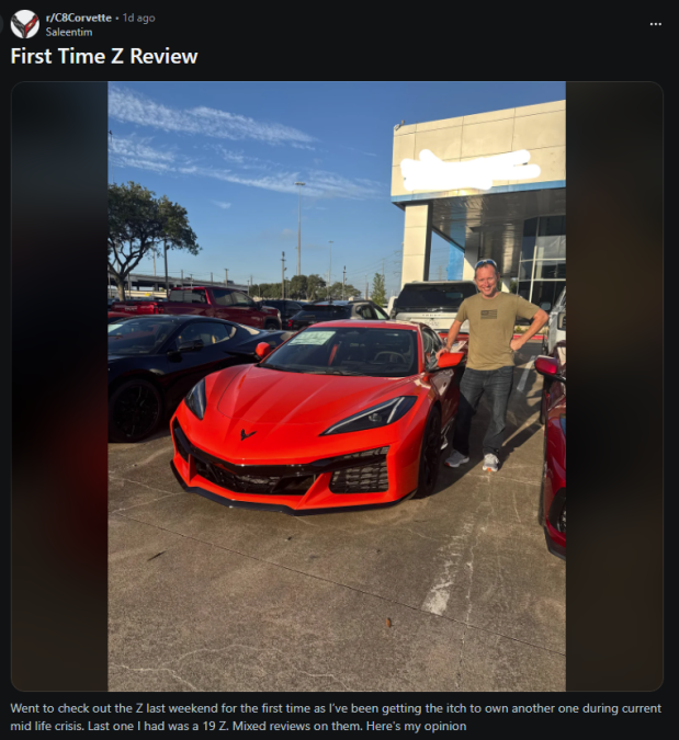 Bright red Chevrolet Corvette sports car parked at a dealership under sunlight. A person stands beside the car with one hand on their hip, smiling. Multiple cars, a dealership building, and a clear blue sky appear in the background.