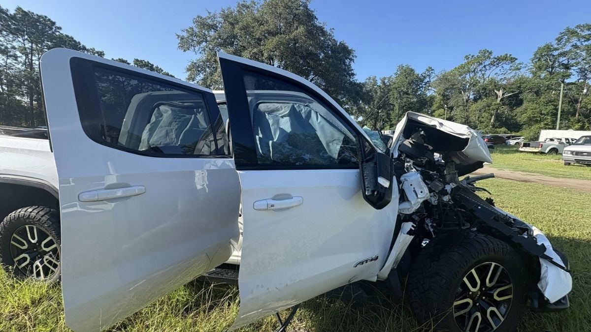 Damaged GMC Sierra 1500 AT4 with a shattered front, crumpled hood, and deployed airbags. The windshield is smashed. Scene conveys an accident's aftermath.