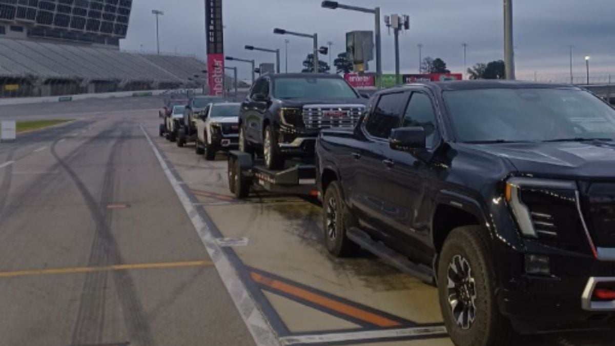 A row of black GMC Sierra EV's on a racetrack, each towing another vehicle under a cloudy sky. The scene conveys anticipation and readiness.