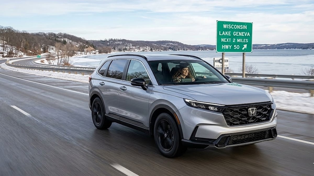 Silver 2026 Honda CR-V Sport-L hybrid cruises past Lake Geneva, Wisconsin highway signs, showcasing its winter-ready design and refined performance