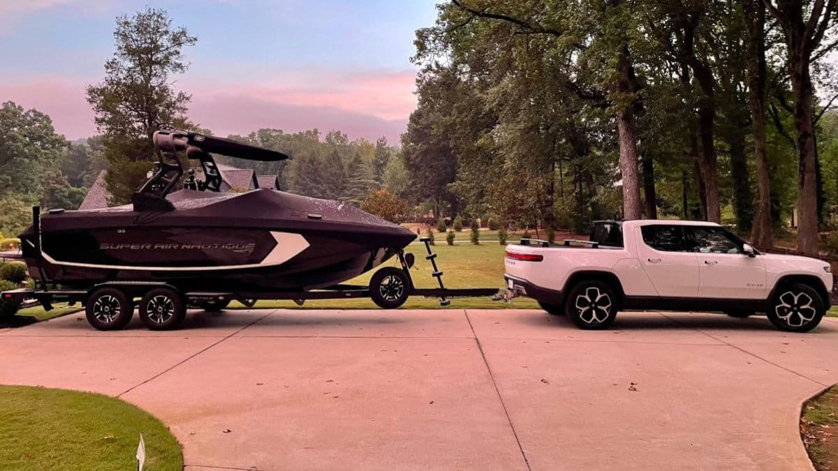 Spencer's white Rivian R1T is parked on a driveway, towing a black speedboat. The scene is set in a lush, green wooded area at sunset, creating a serene and adventurous vibe..