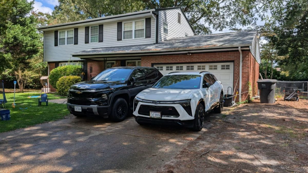 Two 2024 Chevrolet's, one is a Blazer EV and the other is a Silverado EV. They are white and black respecitvely and are parked in a residential driveway showing their front three-quarter angles.