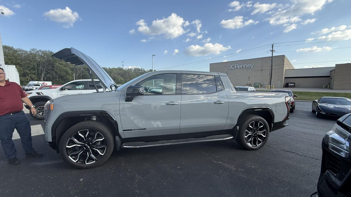 A gray GMC Sierra EV with a modern design and open hood is parked in a lot. The sky is partly cloudy, and a JC Penney store is visible in the background.