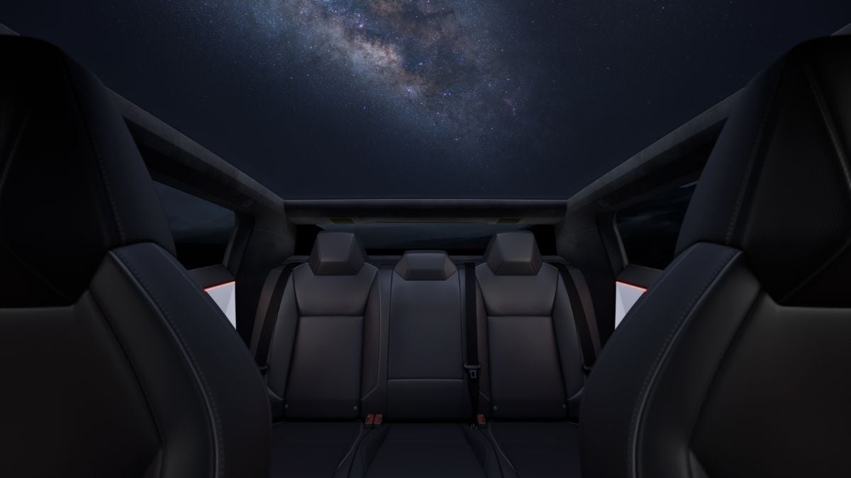 Interior of a Tesla Cybertruck with black seats, viewed from the back. A panoramic glass roof reveals a starry night sky with the Milky Way visible.
