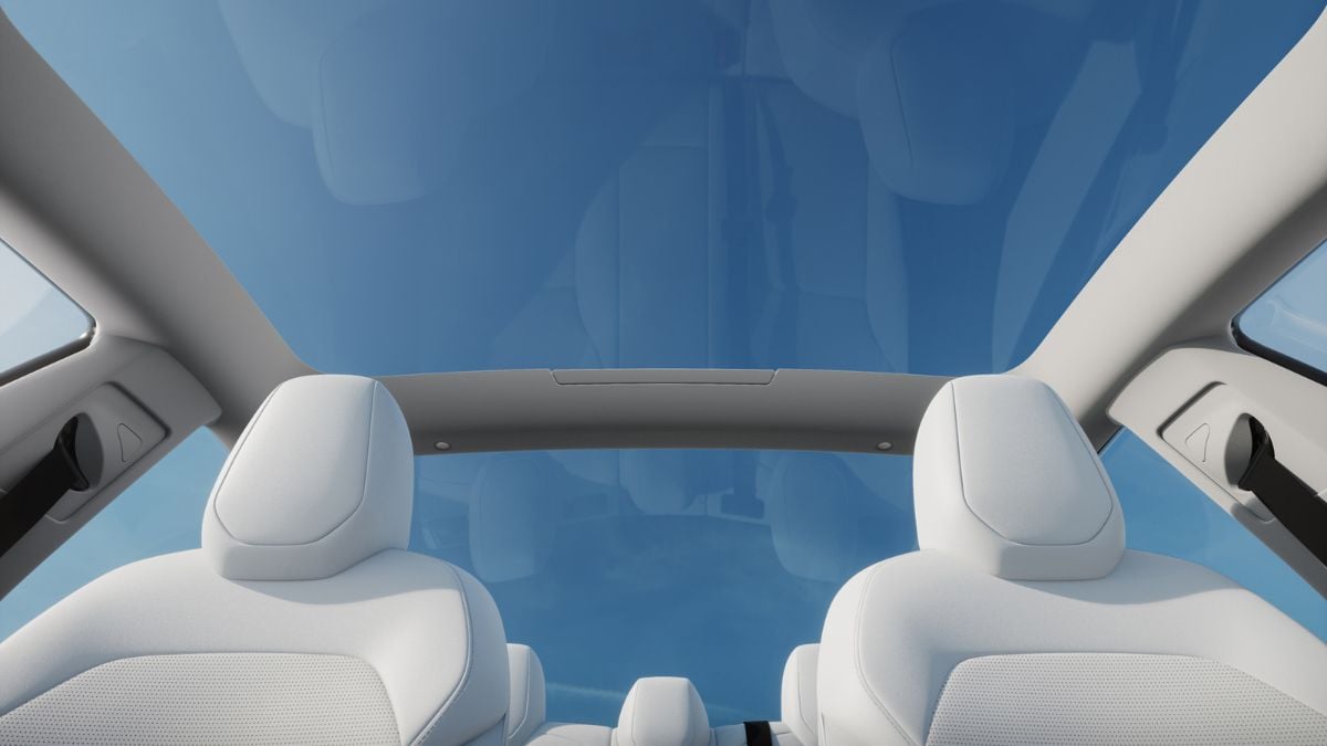 View from inside of a Tesla Model 3 with white seats, looking up at a panoramic glass roof revealing a clear blue sky, conveying a sense of openness and tranquility.
