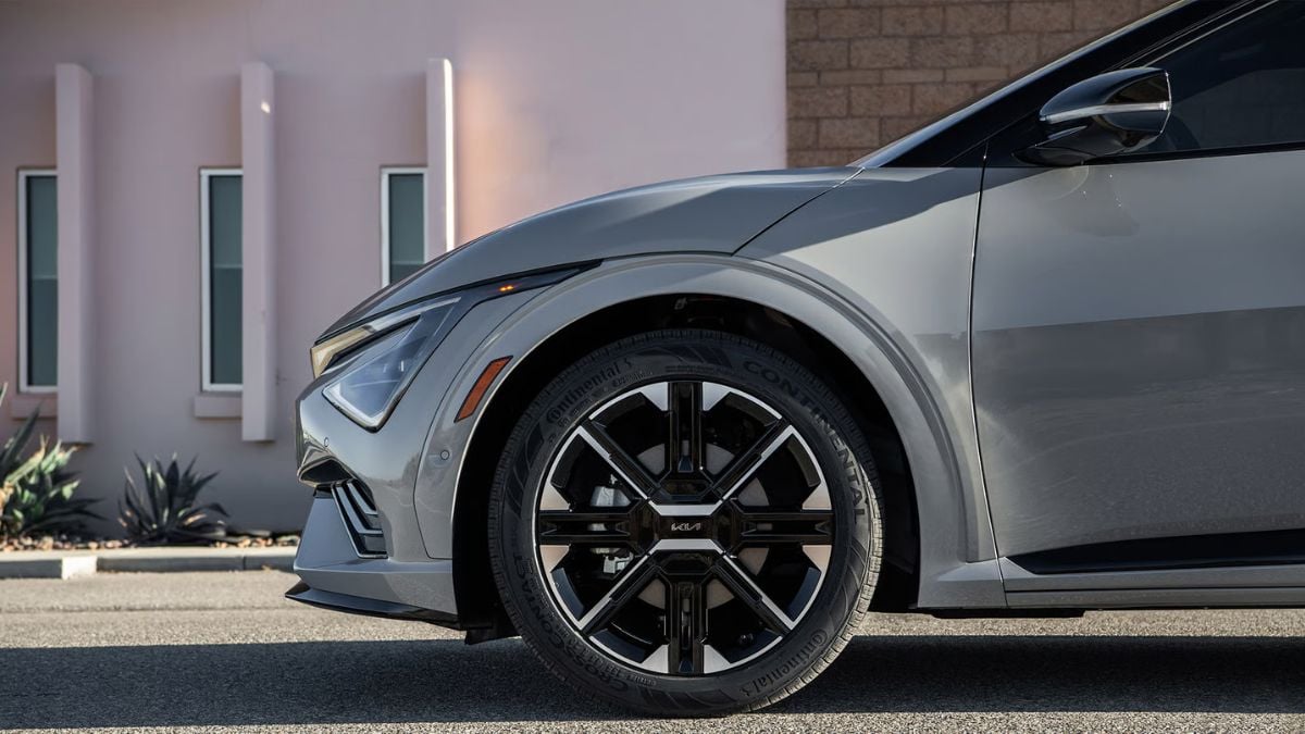 Close-up side view of a silver Kia EV6's front quarter panel, showcasing its distinctive wheel design and modern LED headlight detail against a contemporary architectural background.