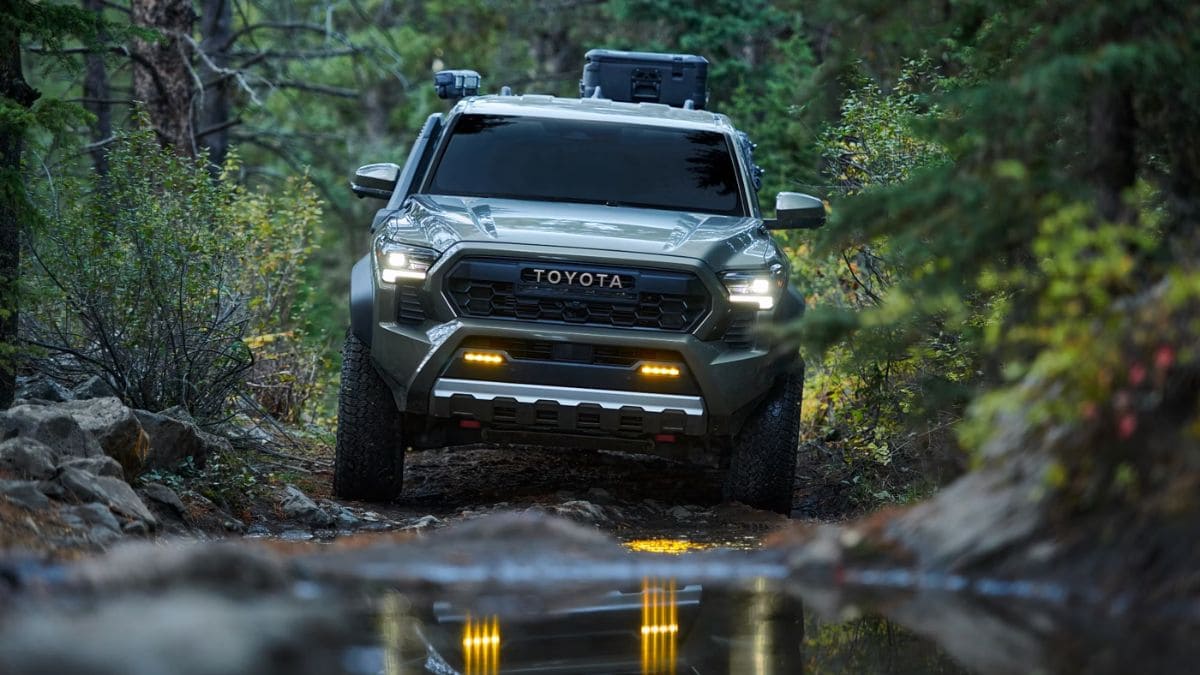 A rugged Toyota Tacoma navigates a forest trail, headlights reflecting in a puddle. Tall trees and dense greenery create an adventurous and off-road atmosphere.