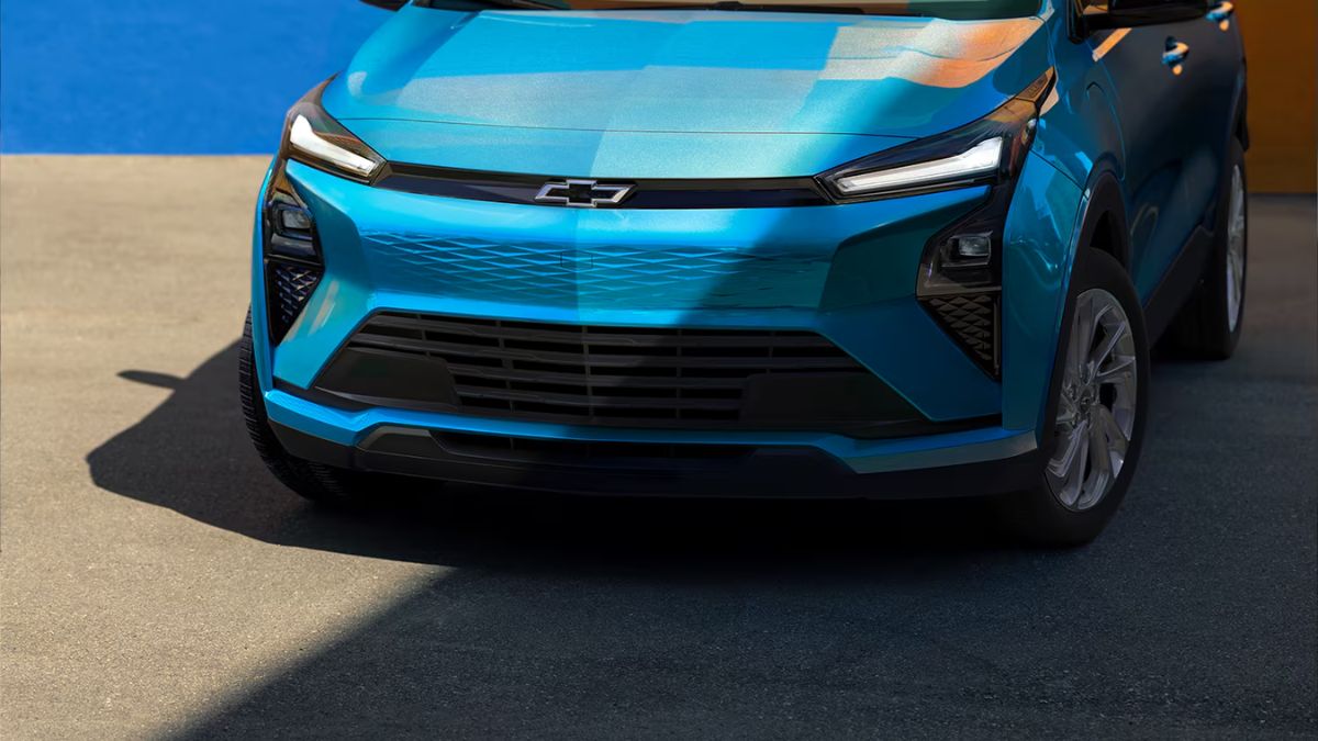 The new 2027 Chevrolet Bolt in bright blue with sleek, angular headlights and a bold front grille. The Chevrolet logo is prominent, and sunlight casts dynamic shadows.