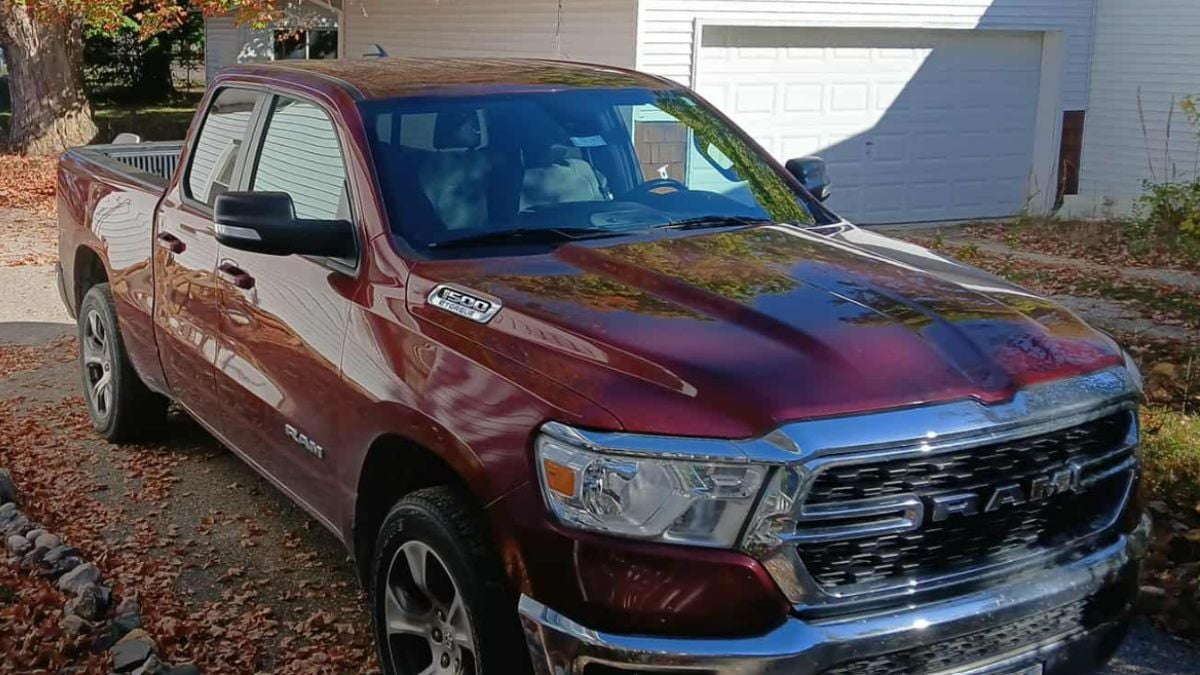 A shiny, burgundy 2022 Ram Bighorn is parked in a sunlit driveway surrounded by autumn leaves. The scene conveys a crisp, fall morning.