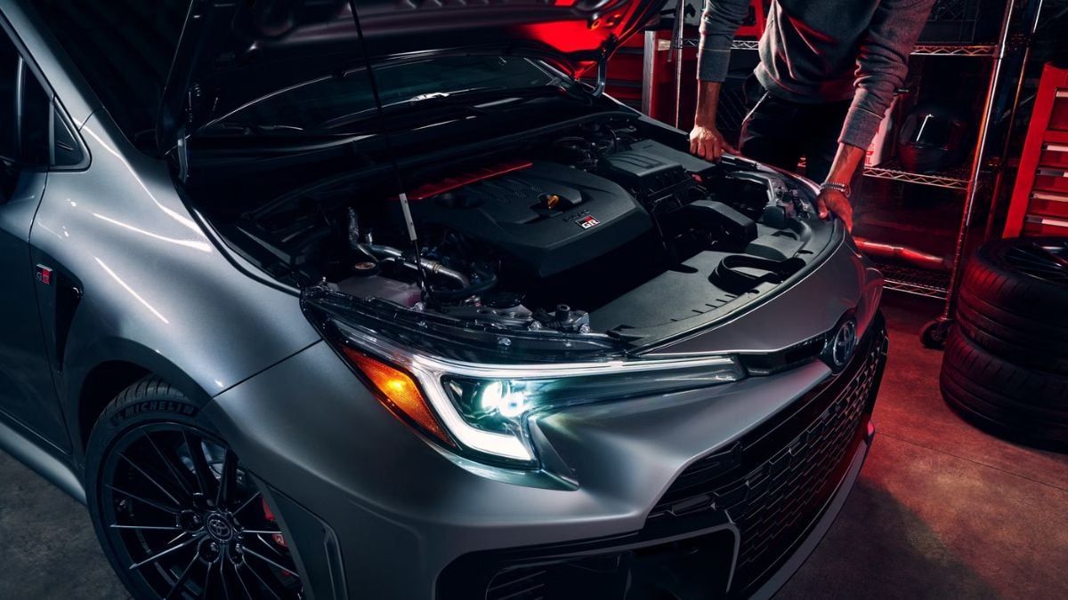 A person examines the engine of a shiny silver Toyota GR Corolla with its hood open in a dimly lit garage, creating a focused and meticulous atmosphere.