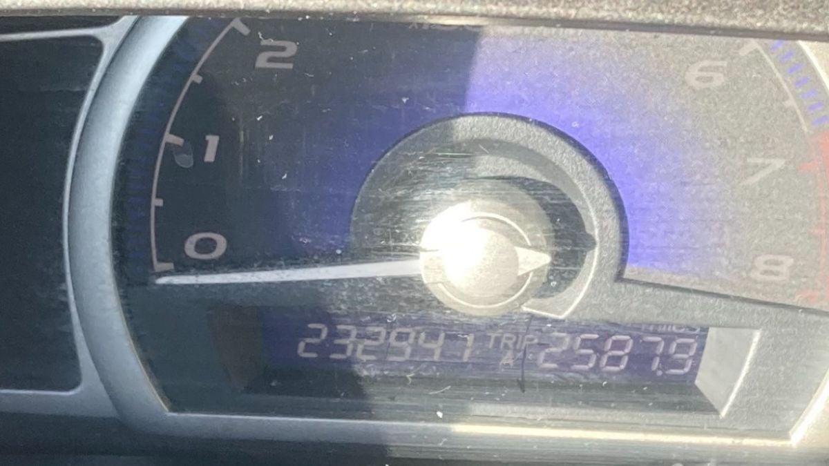 Close-up of Zach's odometer showing 232,947 miles and a trip reading of 258.7 miles. The sun creates a glare on the speedometer display.