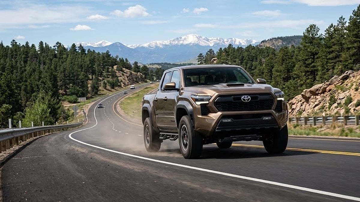A 2025 bronze Toyota Tacoma TRD Pro in motion, climbing a steep highway up Monument Hill, Colorado, with mountains and pine forests