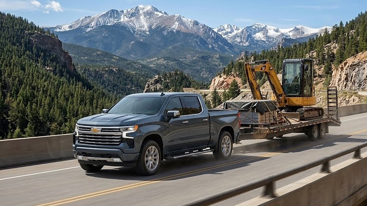 A 2025 Chevy Silverado hauls a heavy equipment trailer up a steep Colorado mountain pass against a scenic alpine backdrop