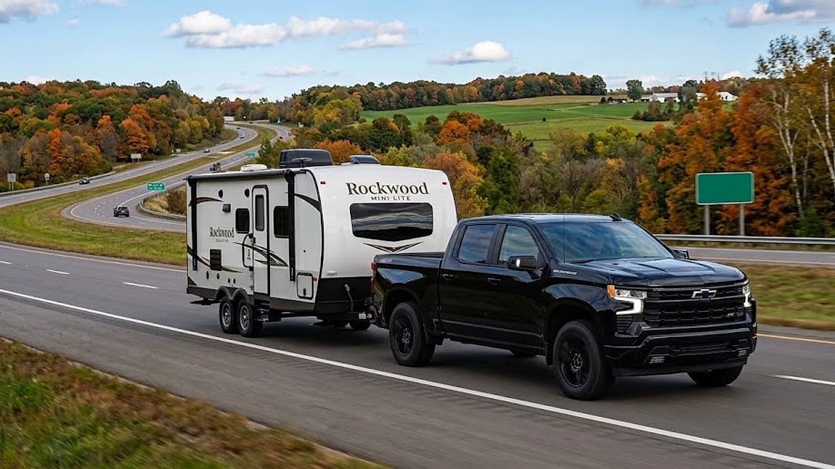 A 2026 Chevy Silverado RST TurboMax pickup trucks pulls a white Rockwood Mini Lite travel trailer along a scenic Wisconsin highway