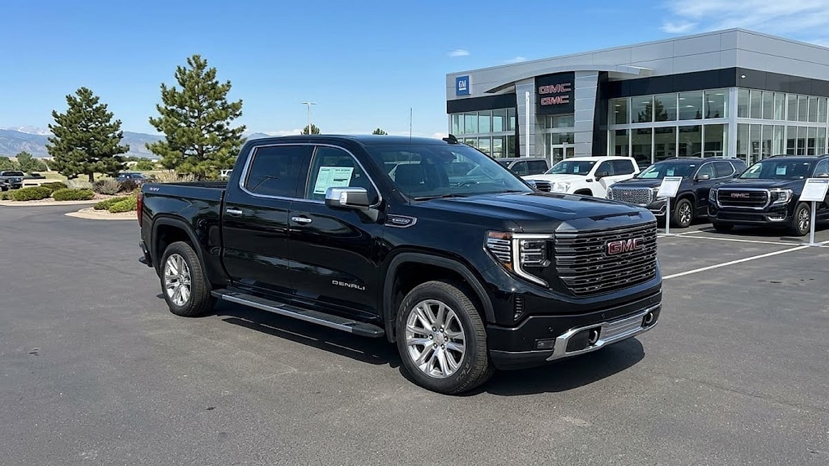 A 2026 GMC Sierra pickup truck is parked among other new vehicles on a modern dealership lot with a mountain range view