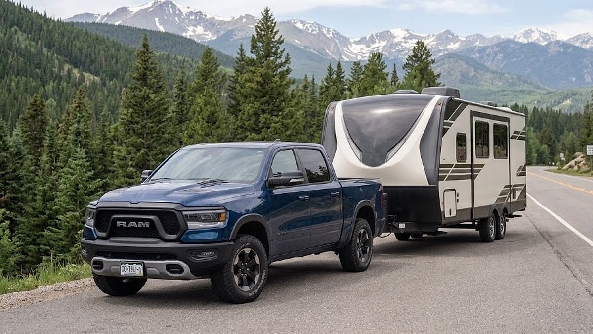 A blue Ram Rebel towing a large fifth-wheel camper on a scenic Colorado mountain highway with snow-capped peaks