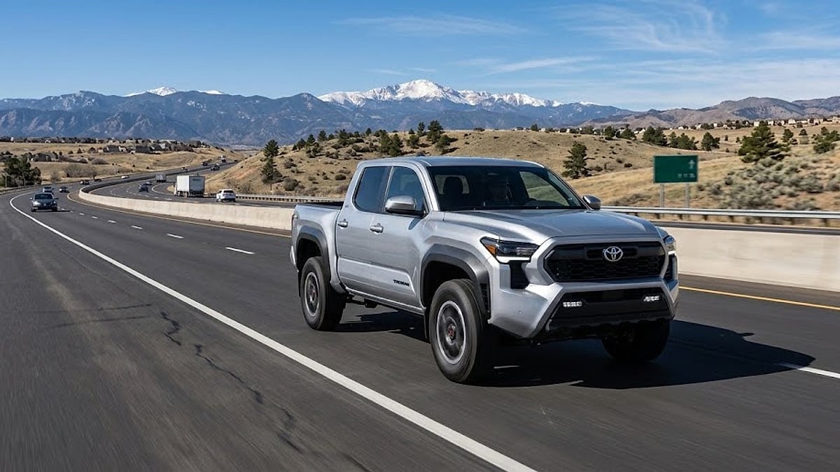 A silver 2024 Toyota Tacoma truck drives on a Colorado highway with snow-capped Rocky Mountains in the background